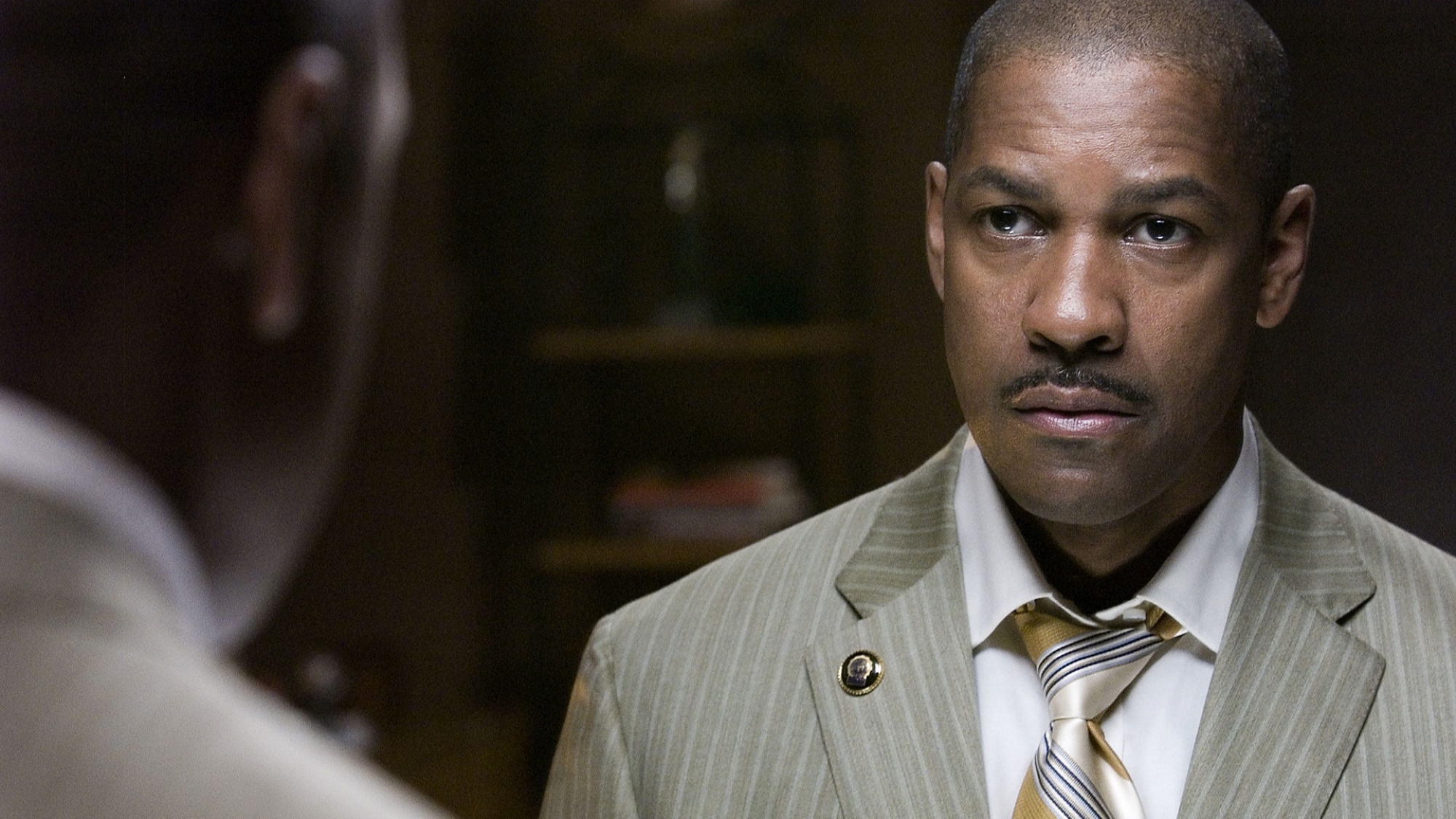 A bald man in a suit stares at a man with his back to the camera.