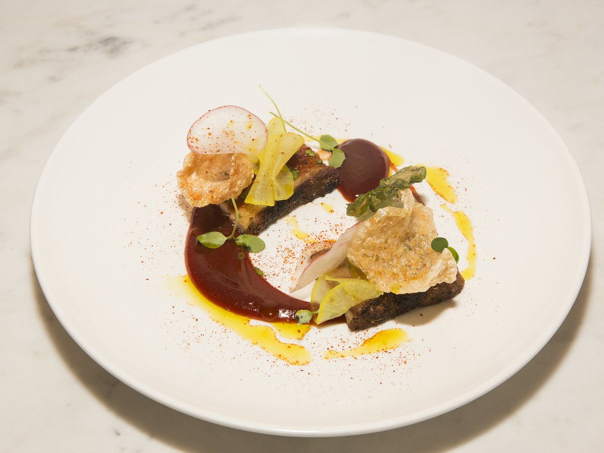 A white plate with two pieces of protein, swooshes of sauce, and delicate garnishes.